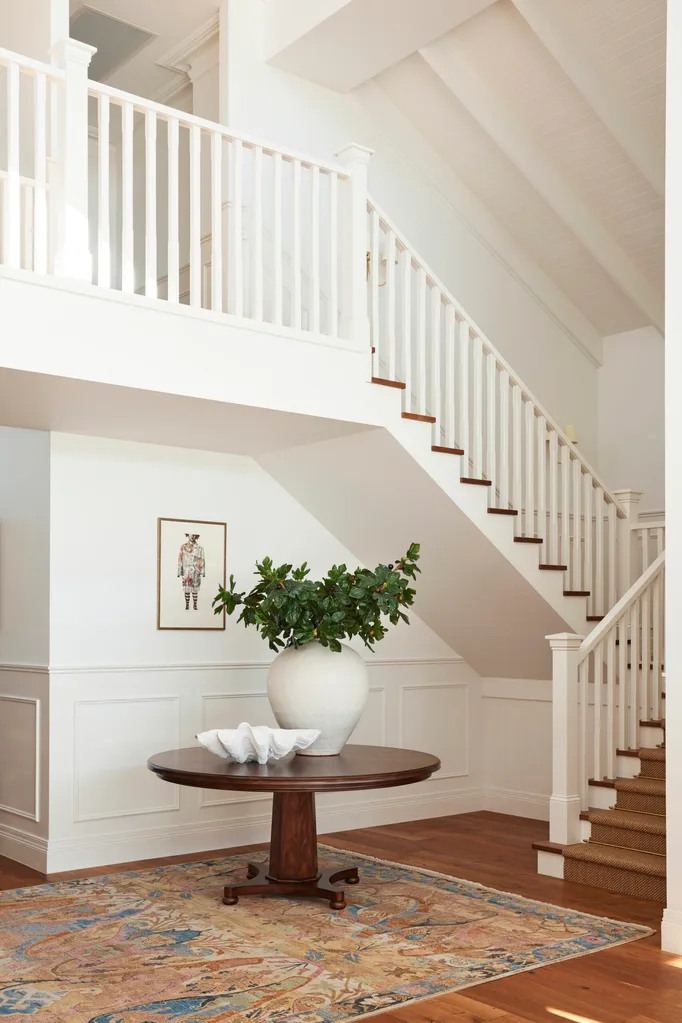 Spacious white foyer with wooden staircase, round table, large vase of greenery, decorative bowl, and patterned rug.