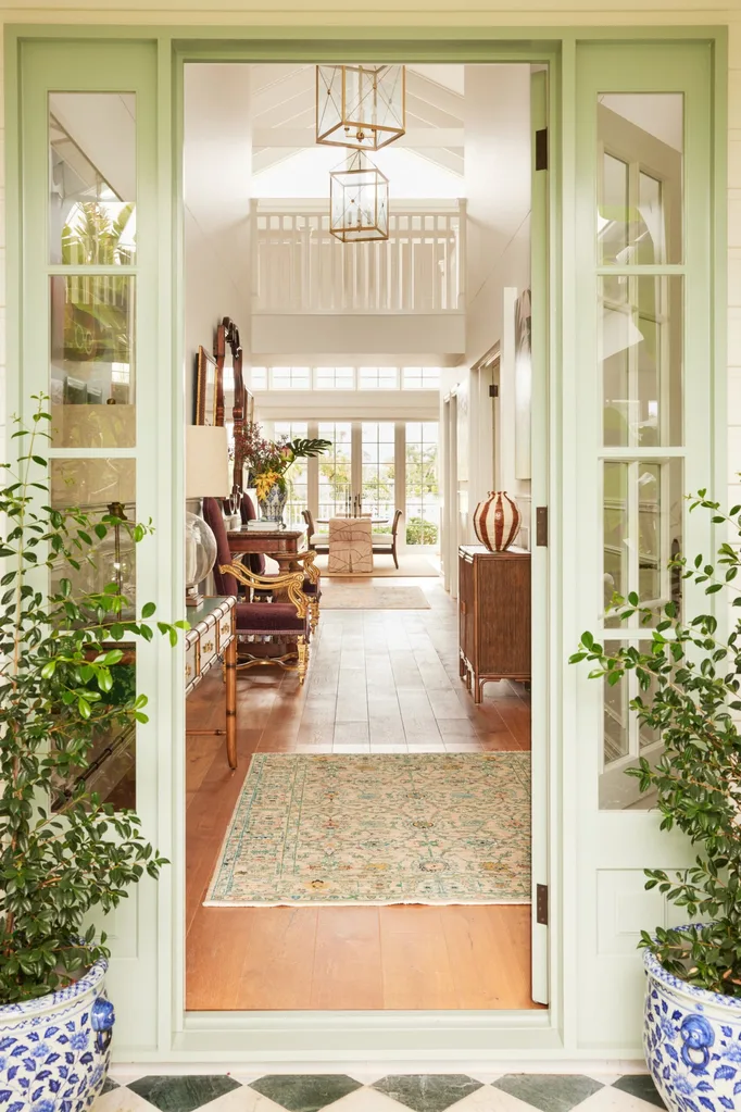 Elegant hallway with light green doors, wooden floor, ornate furniture, plants, and natural light from large windows.