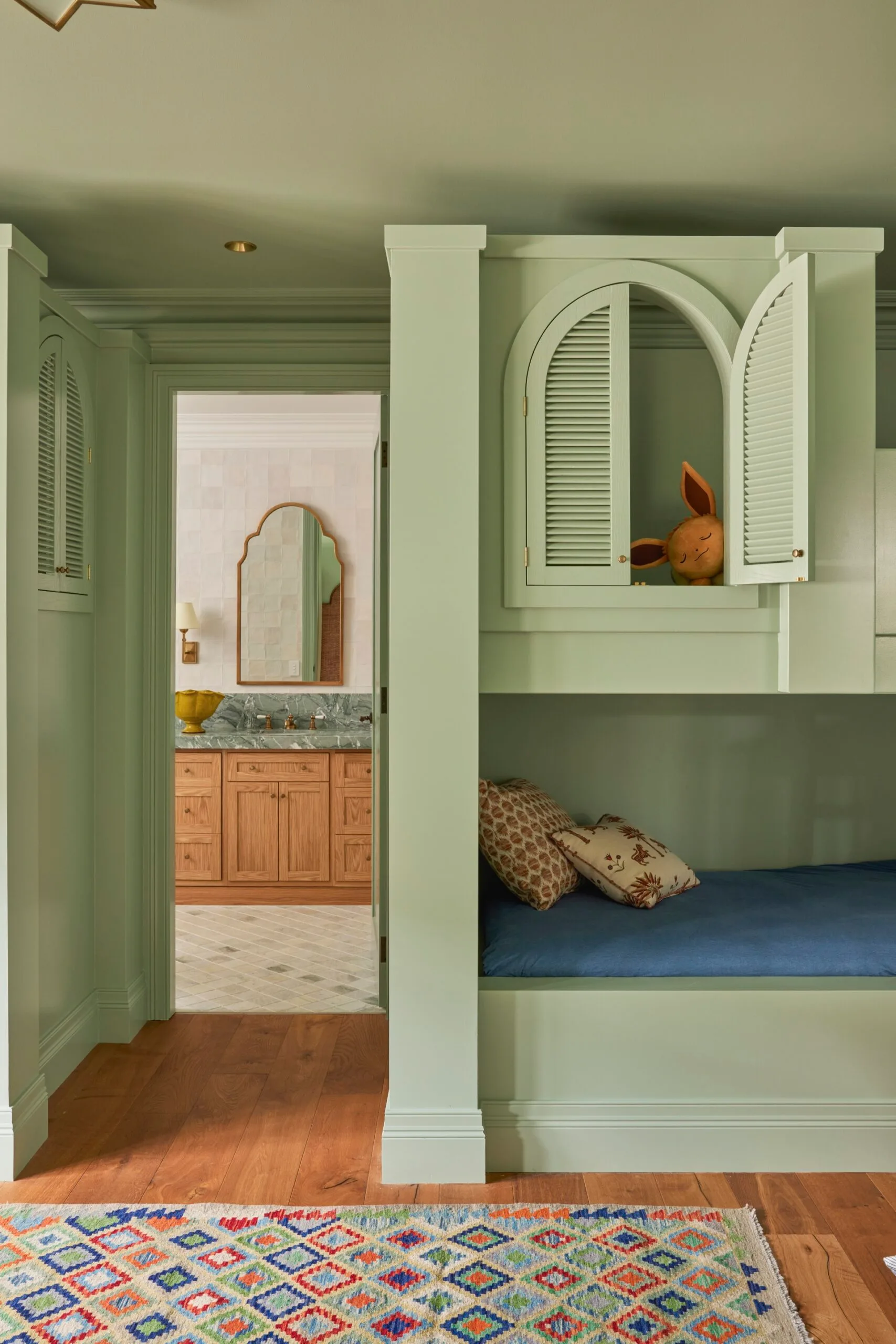 Green bunk bed room with open cabinet; colorful rug and a bathroom with wooden cabinetry in the background.