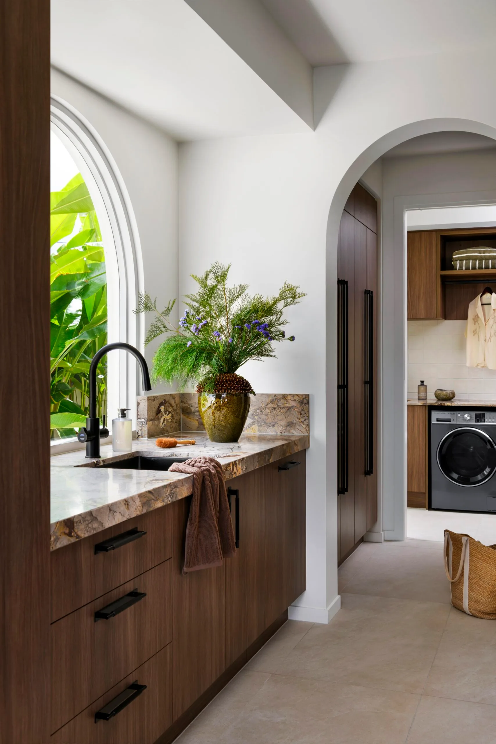 Modern kitchen with wood cabinets, marble countertop, black faucet, and a vase of plants under an arched window.
