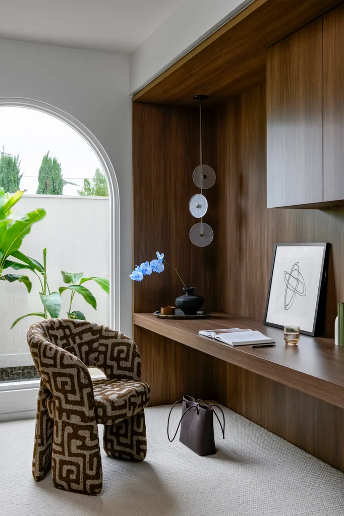 Cozy home office with patterned chair, wooden desk, decorative vase, and abstract artwork near large arched window.