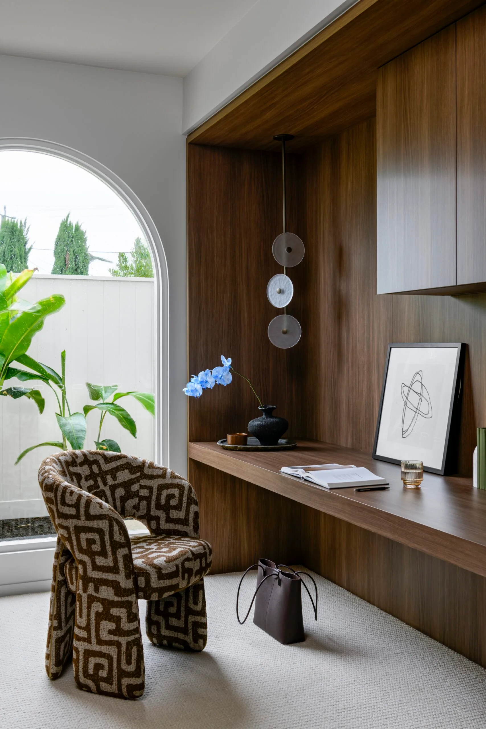 Cozy home office with patterned chair, wooden desk, decorative vase, and abstract artwork near large arched window.