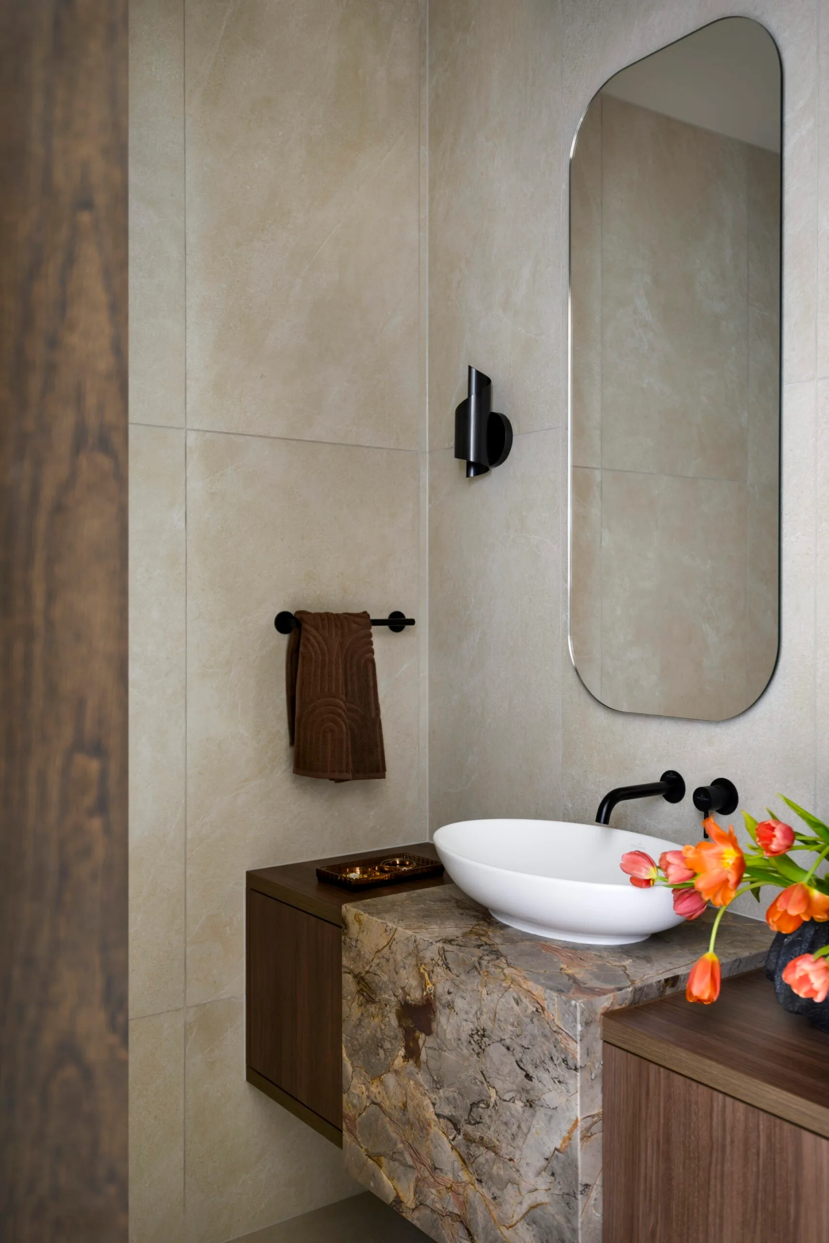 Modern bathroom with beige tiles, stone vanity, oval sink, large mirror, and orange tulips; minimal and elegant design.