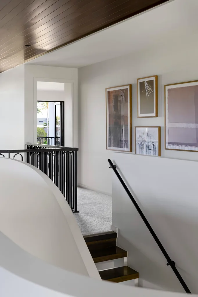 Upper hallway with stairs, black railing, light walls, art on walls, and carpet, leading to sunlit room with window.