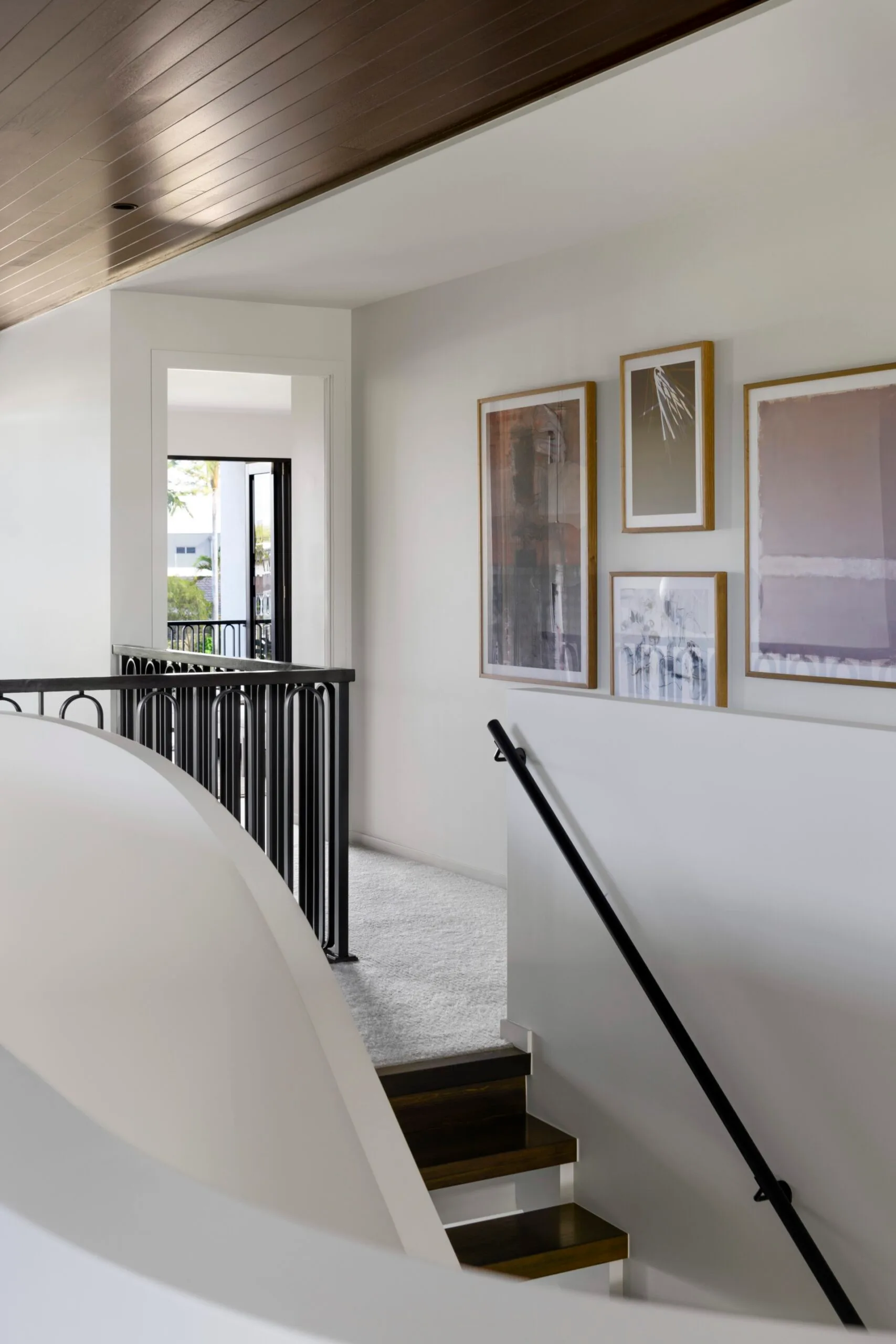 Upper hallway with stairs, black railing, light walls, art on walls, and carpet, leading to sunlit room with window.