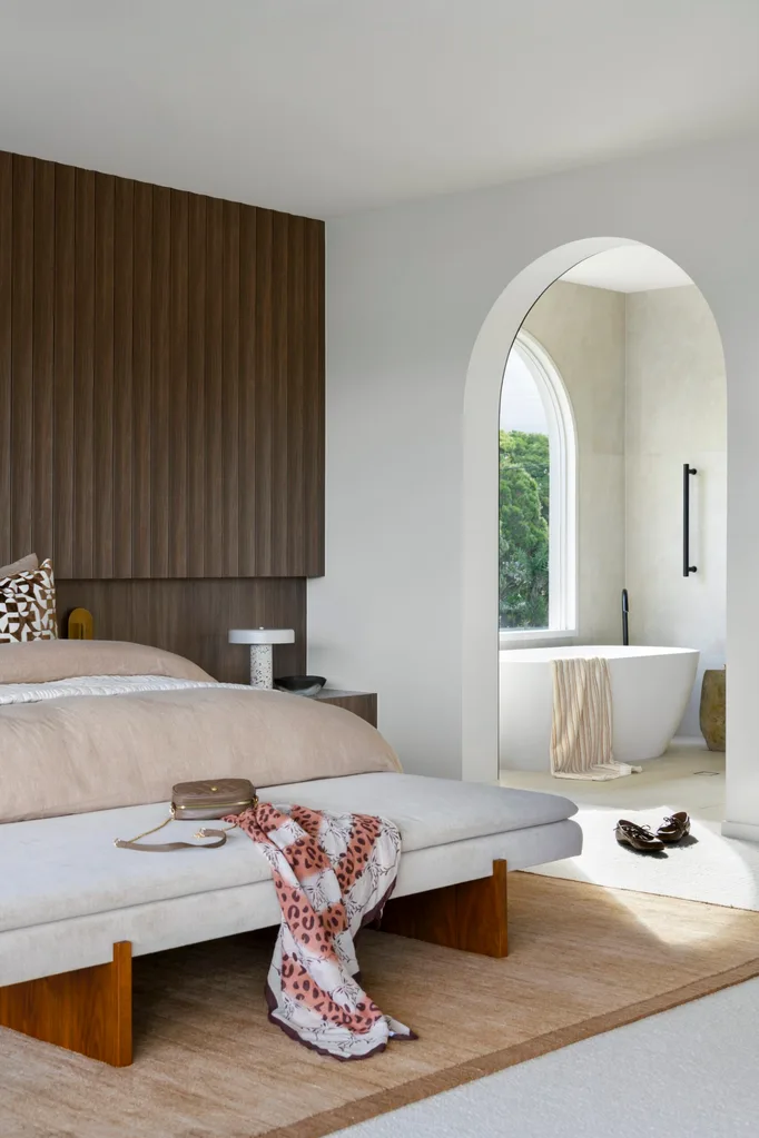Modern bedroom with a wooden accent wall, a plush bed, bench with a scarf, and an archway leading to a bathroom with a bathtub.