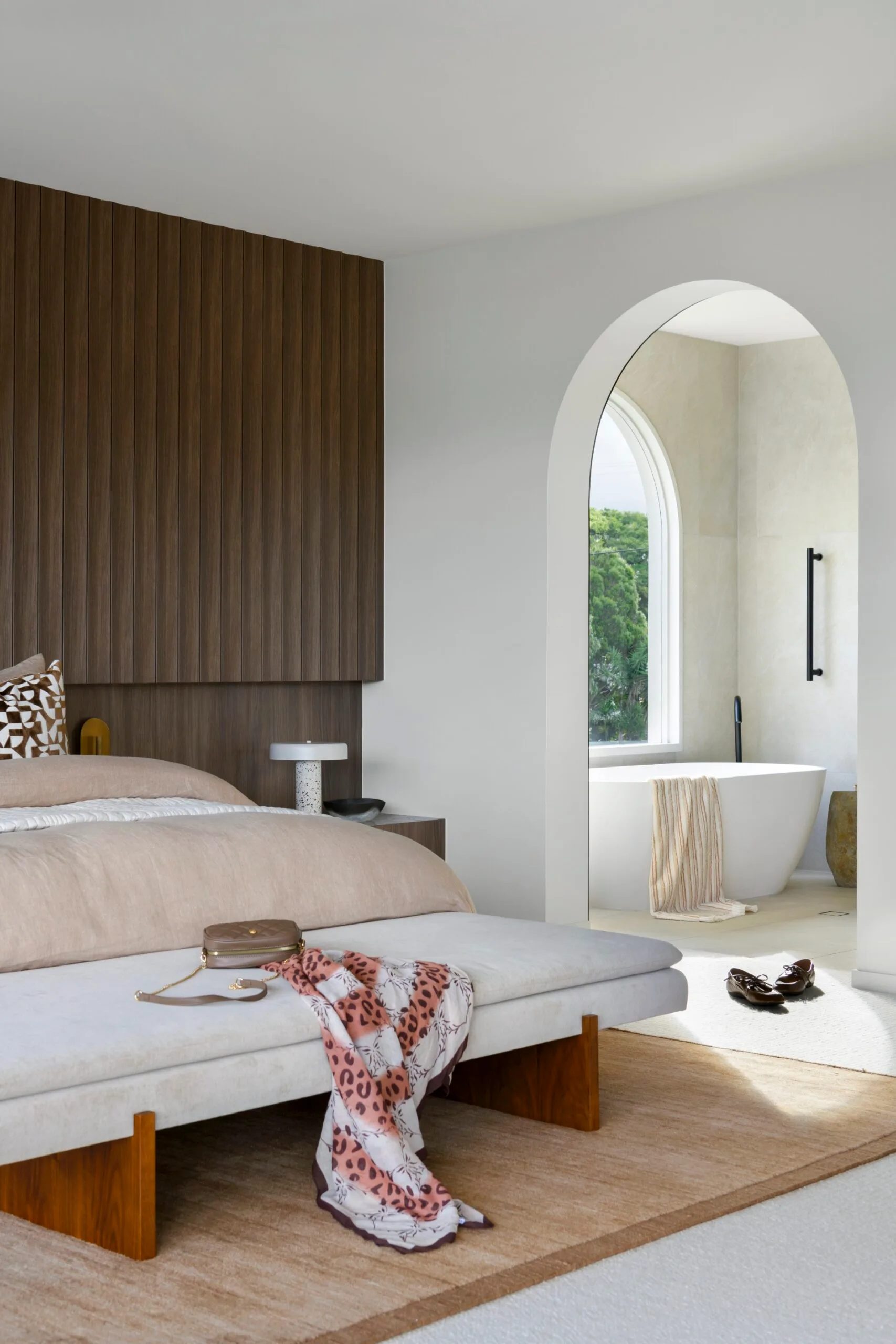 Modern bedroom with a wooden accent wall, a plush bed, bench with a scarf, and an archway leading to a bathroom with a bathtub.