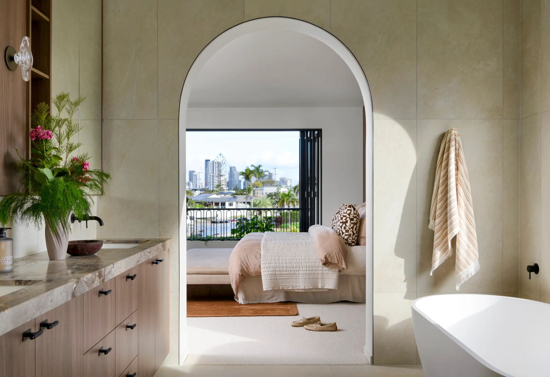 Modern bathroom with archway view to bedroom, city skyline visible. Natural light illuminates plants and decor.