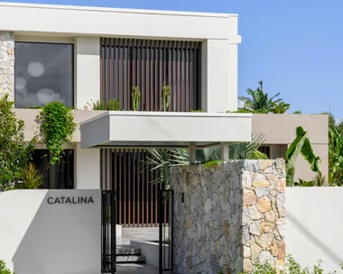 Modern two-story building with "CATALINA" sign, stone entrance, and lush greenery under a clear blue sky.