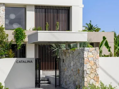 Modern two-story building with "CATALINA" sign, stone entrance, and lush greenery under a clear blue sky.