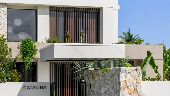 Modern two-story building with "CATALINA" sign, stone entrance, and lush greenery under a clear blue sky.