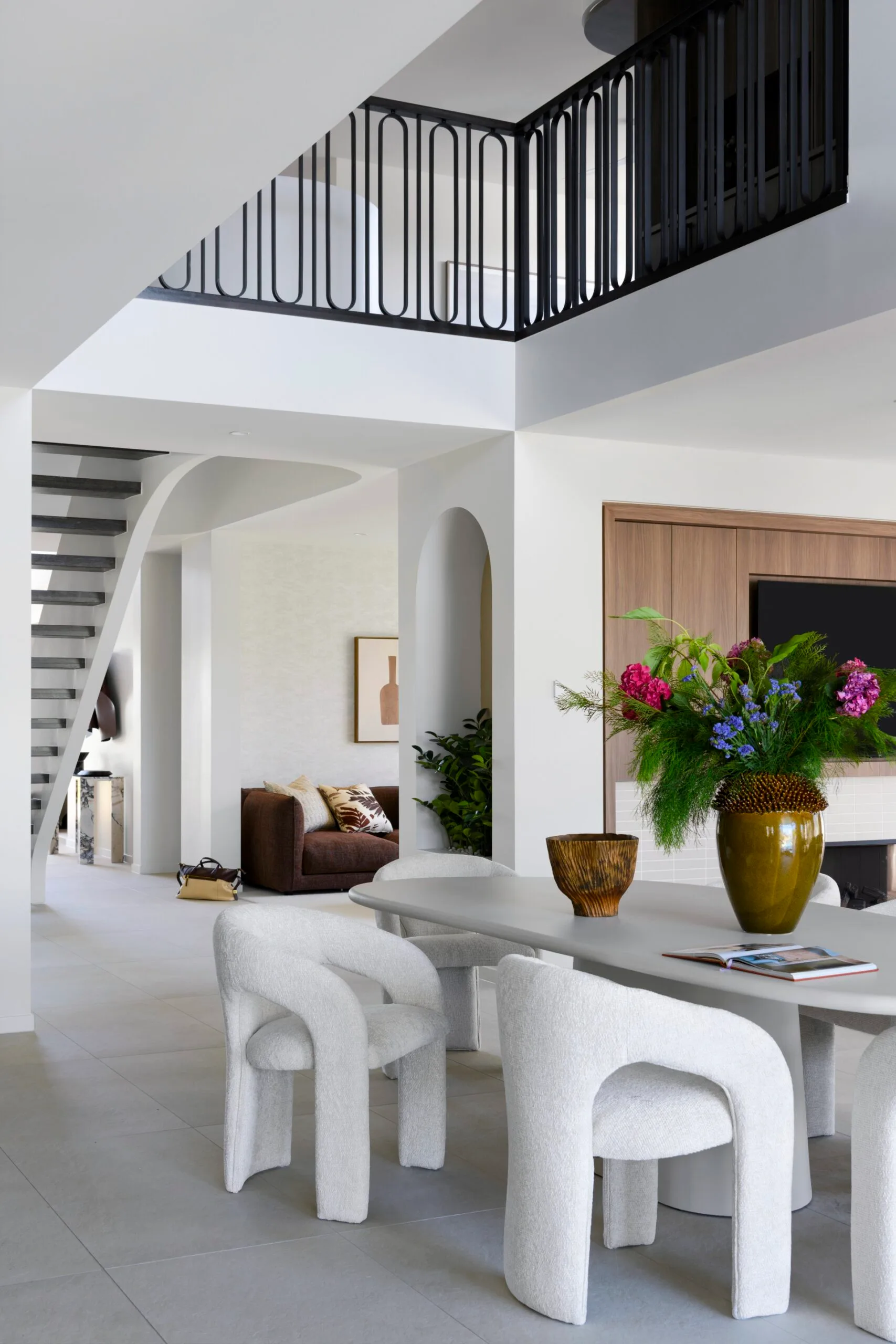 Modern living room with white chairs, a gray table, staircase, and vibrant floral centerpiece in a stylish interior.