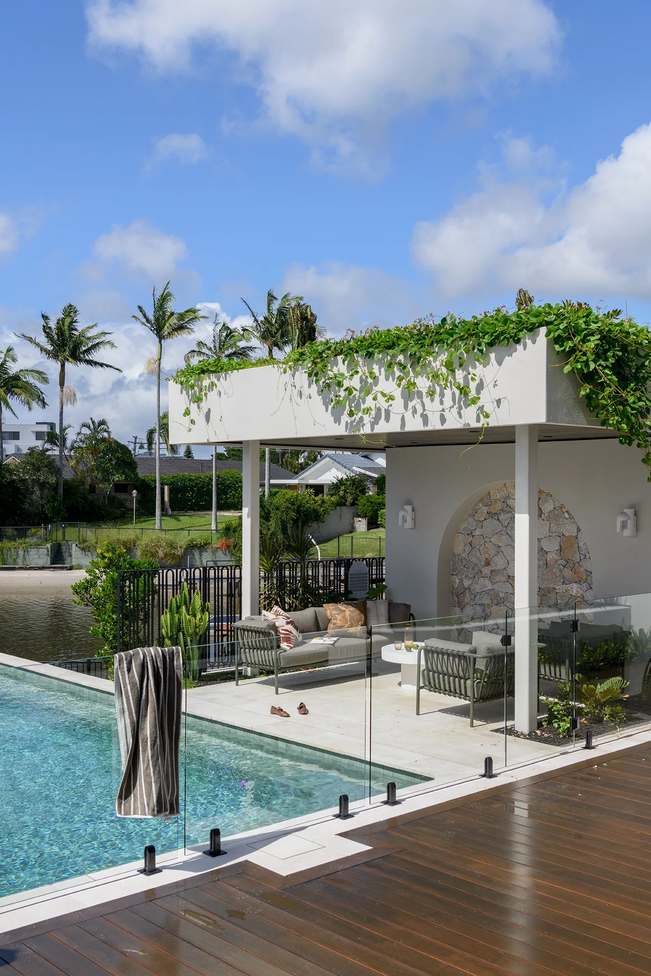 Outdoor pool area with deck, glass fence, and shaded seating under a pergola with green plants, near palm trees.