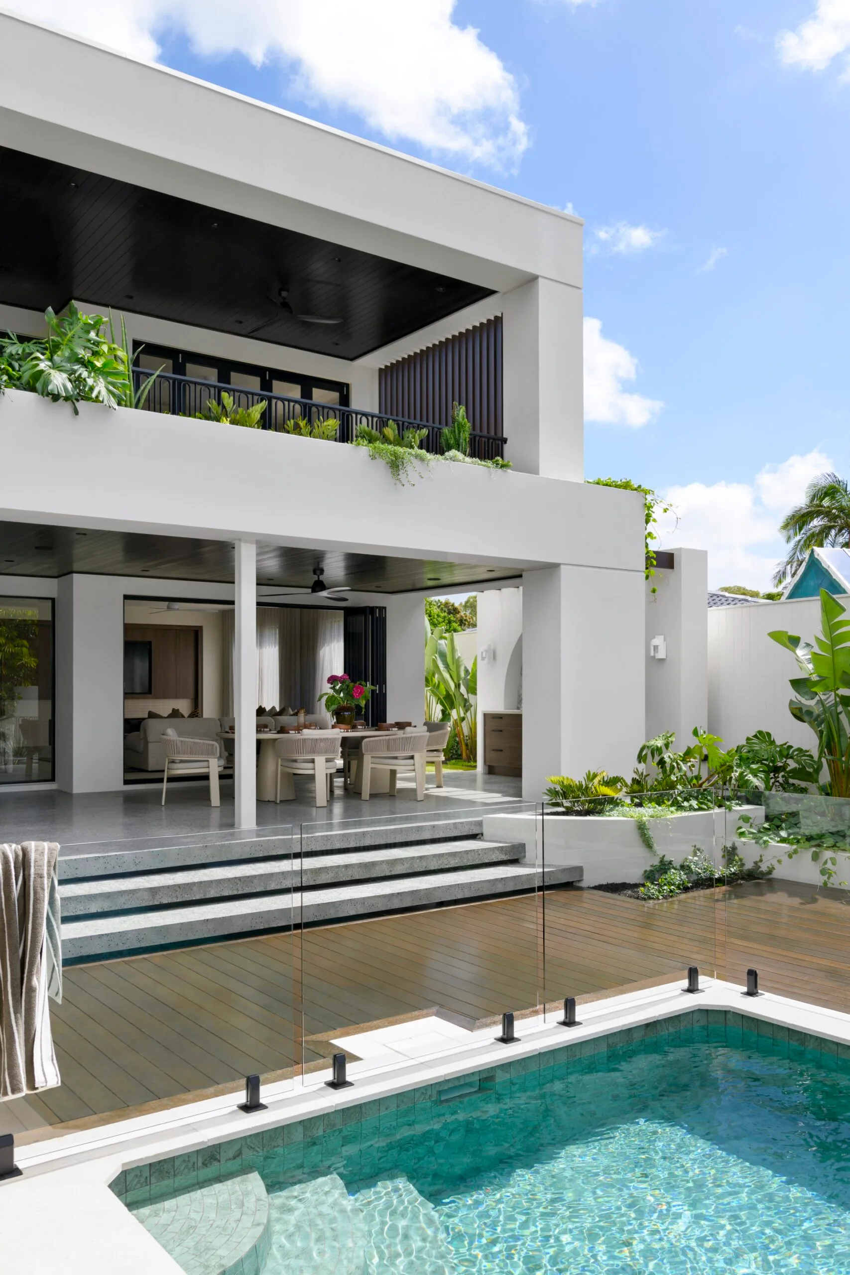 Modern two-story house with outdoor dining area, balcony, and a pool, surrounded by lush greenery.