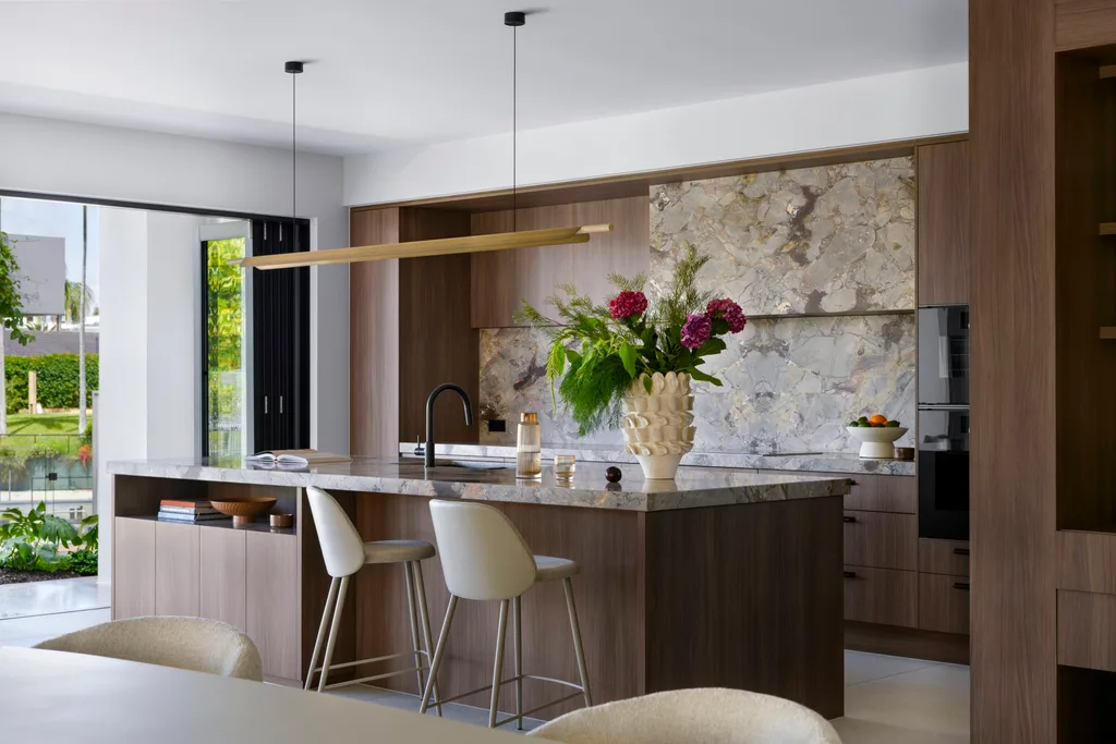 Modern kitchen with marble island, wooden cabinets, decorative vase with flowers, and two bar stools under pendant light.