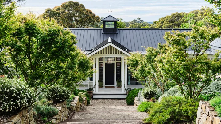 Pathway leads to a charming house with a black roof, surrounded by lush greenery and stone walls, under a partly cloudy sky.