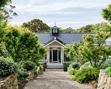 Pathway leads to a charming house with a black roof, surrounded by lush greenery and stone walls, under a partly cloudy sky.