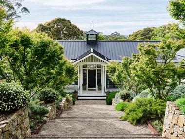Pathway leads to a charming house with a black roof, surrounded by lush greenery and stone walls, under a partly cloudy sky.