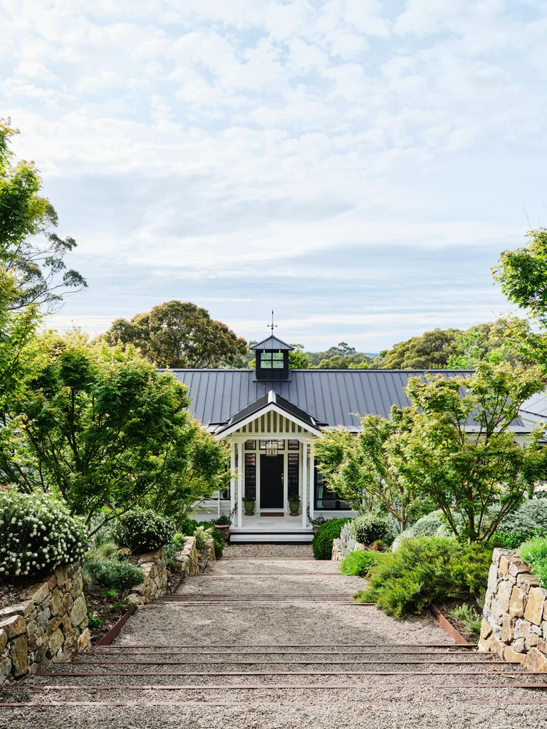 Pathway leads to a charming house with a black roof, surrounded by lush greenery and stone walls, under a partly cloudy sky.