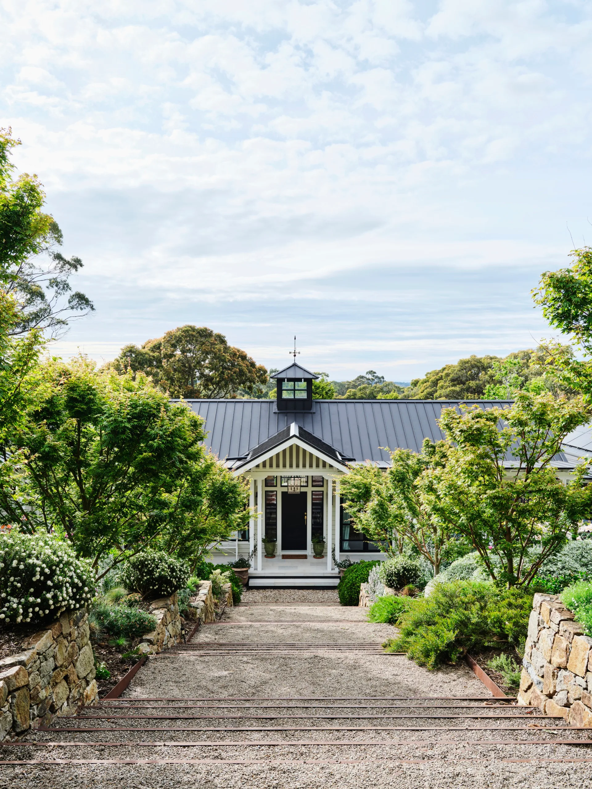Pathway leads to a charming house with a black roof, surrounded by lush greenery and stone walls, under a partly cloudy sky.