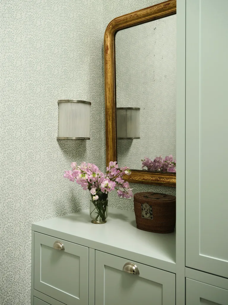 Floral arrangement on a cabinet with a wicker basket, ornate mirror, and textured wallpaper in a cozy setting.