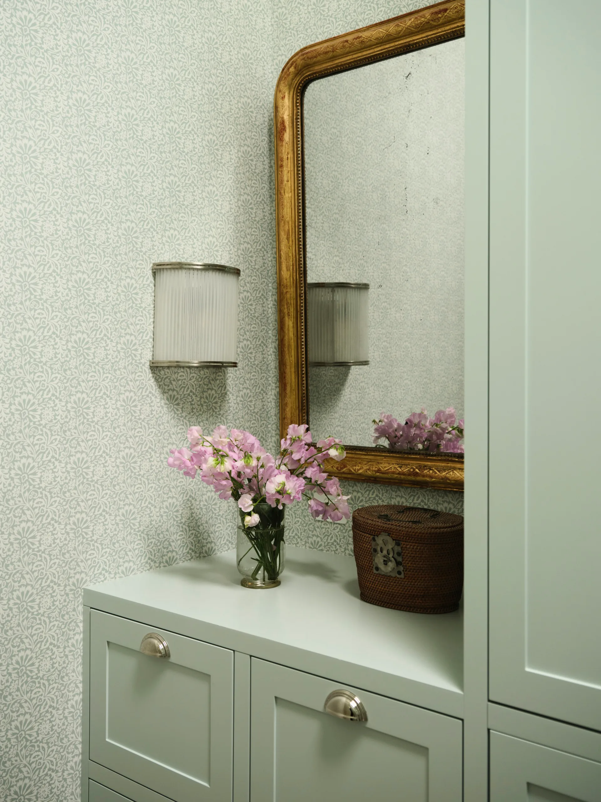 Floral arrangement on a cabinet with a wicker basket, ornate mirror, and textured wallpaper in a cozy setting.