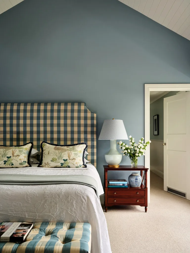 Elegant bedroom with a checkered headboard, floral pillows, side table with vase and lamp, and a blue accent wall.