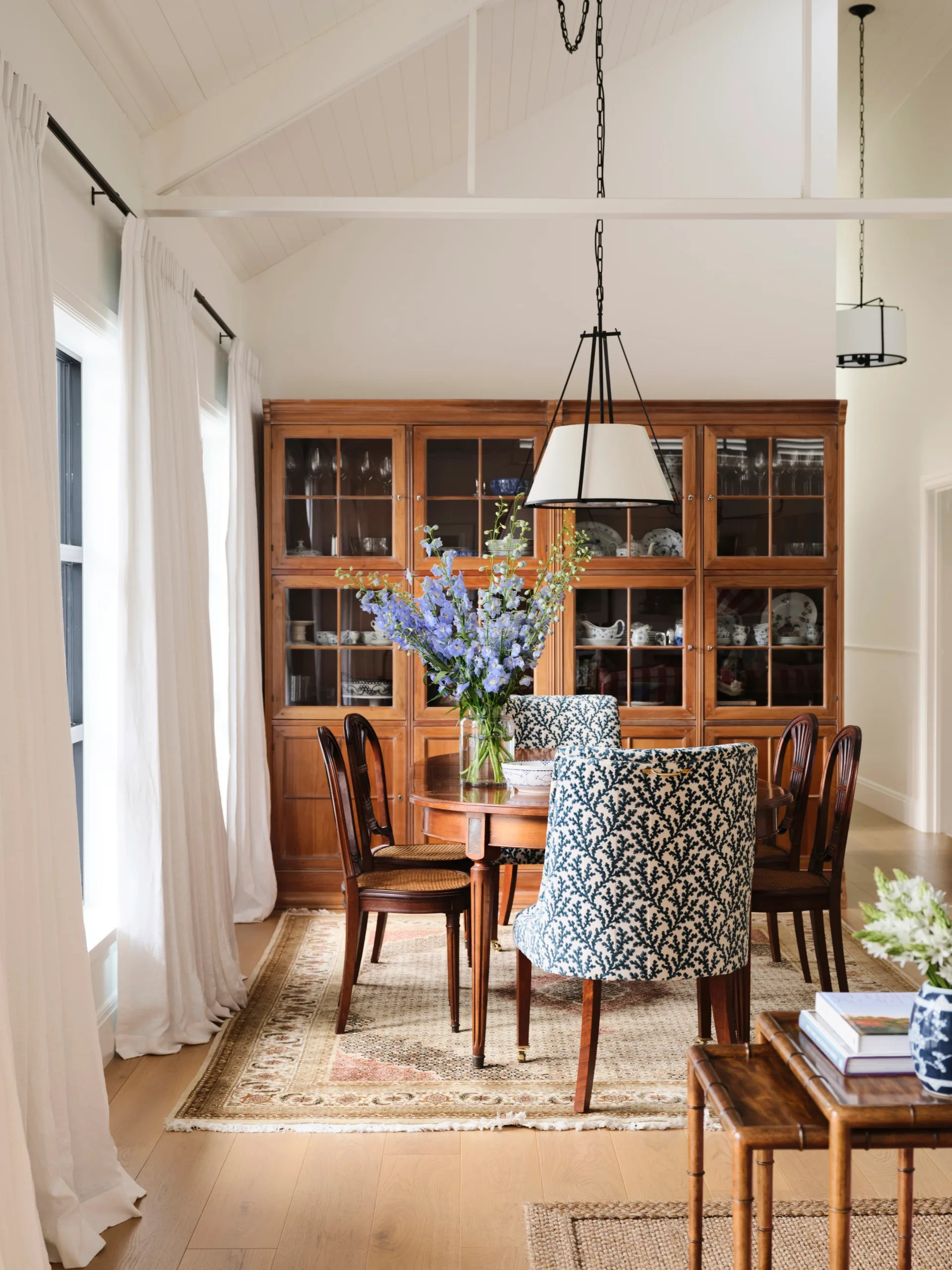Dining room with a wooden table, blue patterned chairs, wooden cabinet, and vase with purple flowers under a pendant light.