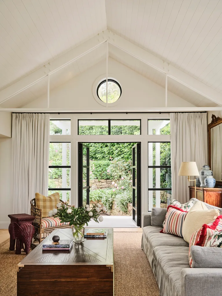 Living room with vaulted ceiling, striped furniture, glass doors open to garden, and a wooden coffee table with flowers.