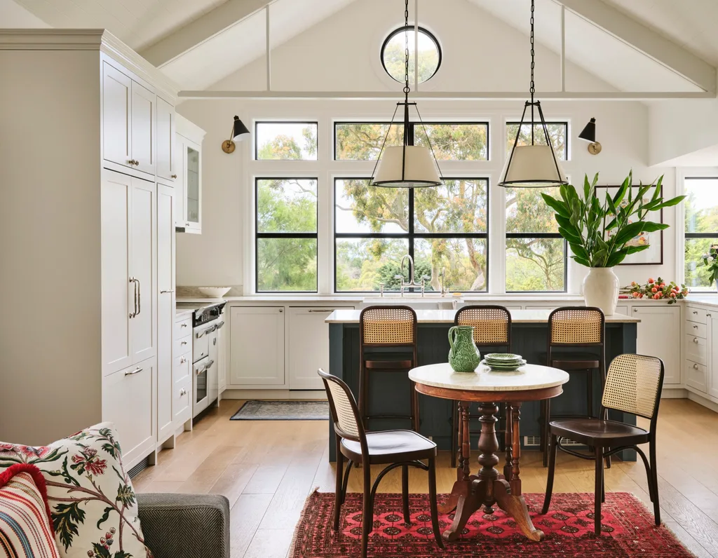 Bright kitchen with white cabinets, a round wooden table, wicker chairs, large windows, and green plants.