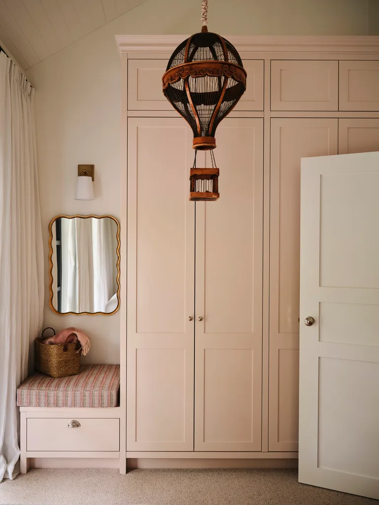 Wardrobe with light pink doors, hot air balloon pendant, mirror, cushioned bench with basket, and curtain.