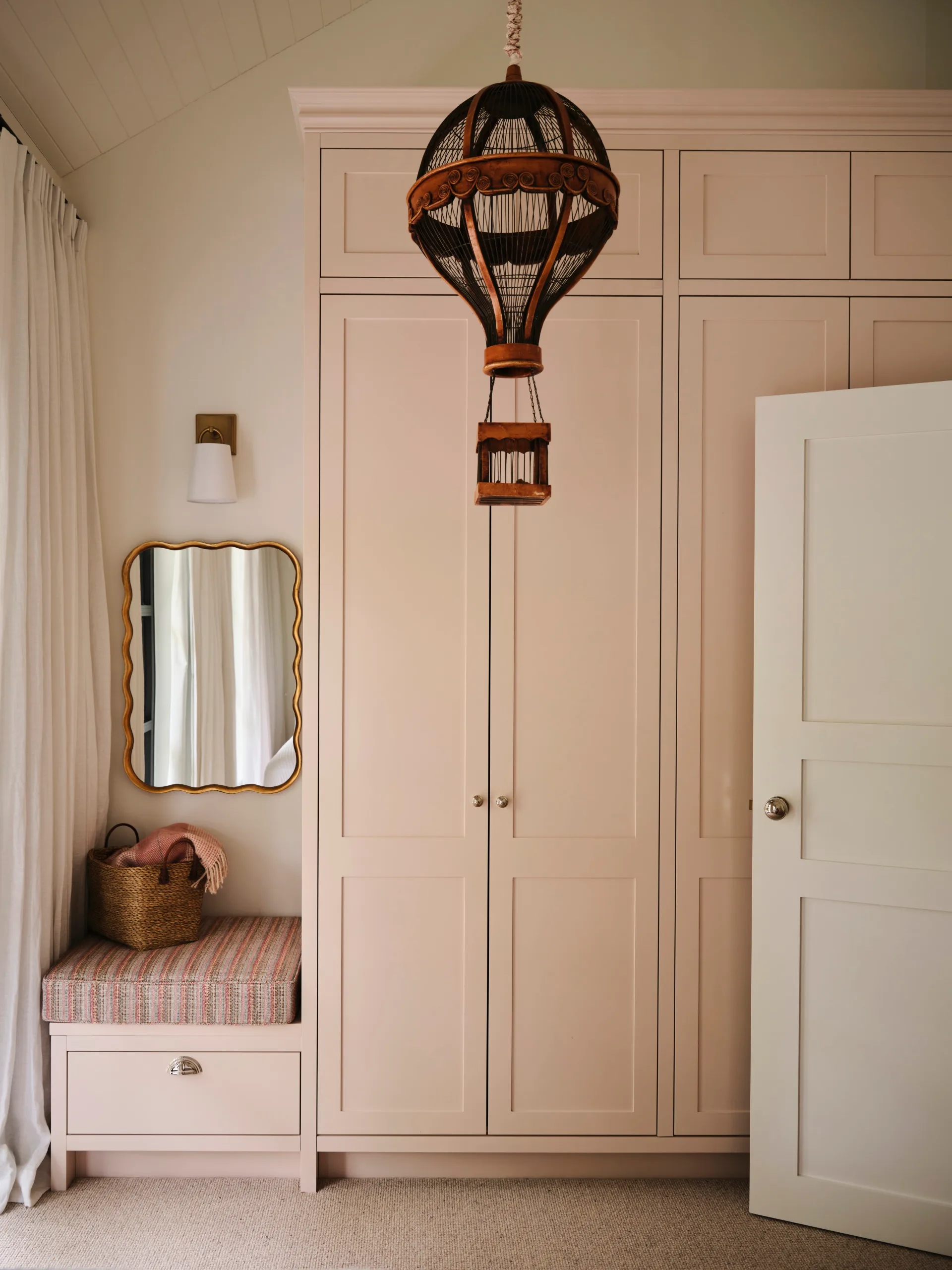 Wardrobe with light pink doors, hot air balloon pendant, mirror, cushioned bench with basket, and curtain.