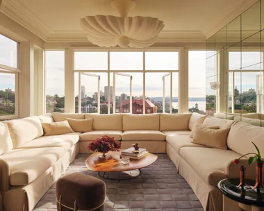 Cream sectional sofa in bright living room with cityscape views through north-facing windows, a wooden coffee table, and decorative ceiling light.