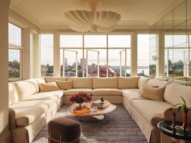 Cream sectional sofa in bright living room with cityscape views through north-facing windows, a wooden coffee table, and decorative ceiling light.