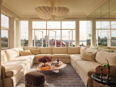 Cream sectional sofa in bright living room with cityscape view, wooden coffee table, and decorative ceiling light.