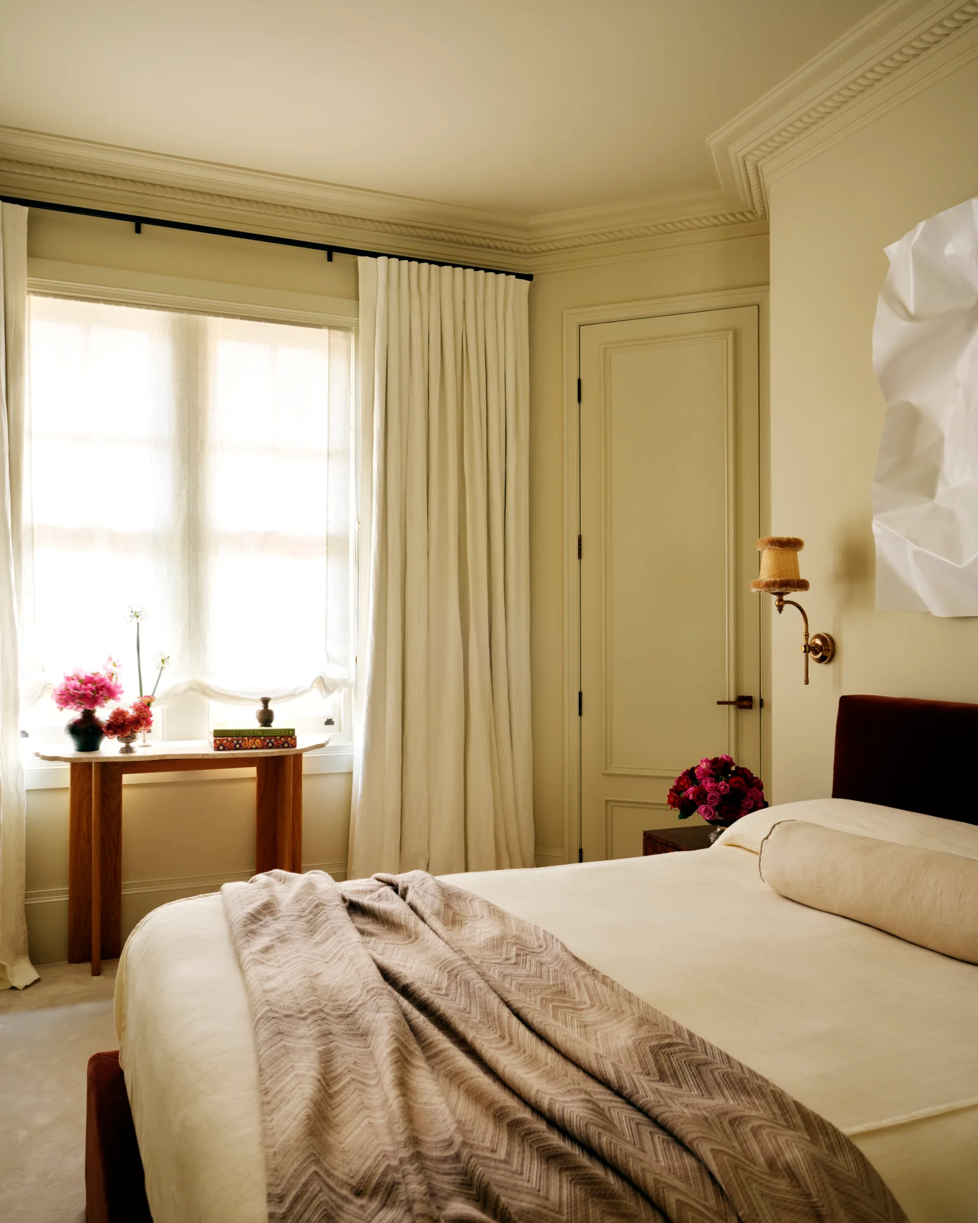 Cozy bedroom with a cream color palette, featuring a bed, beige blanket, soft curtains, and wall art beside a wooden table.