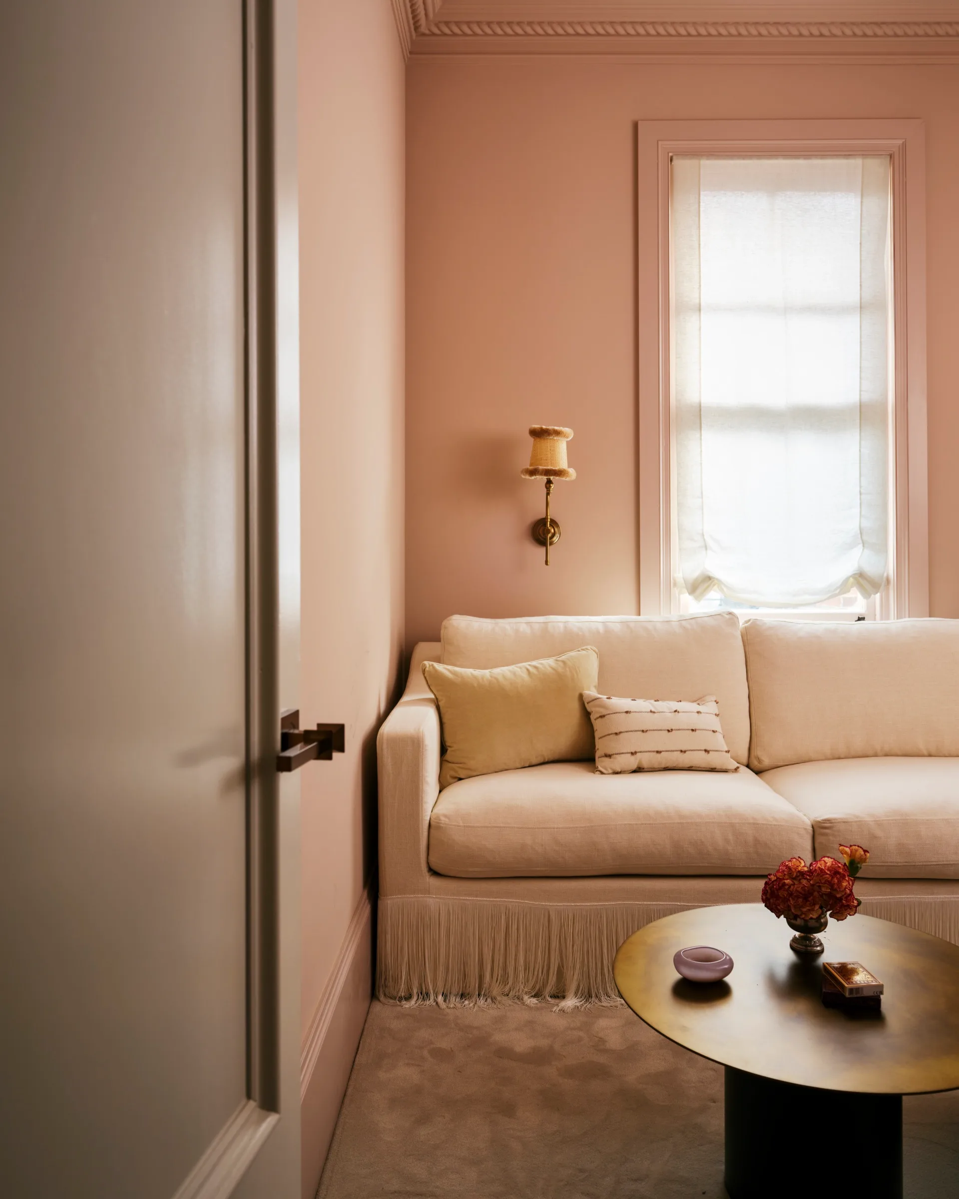 Cozy beige living room with a sofa, pillows, a wall sconce, and a round coffee table with flowers and decor.