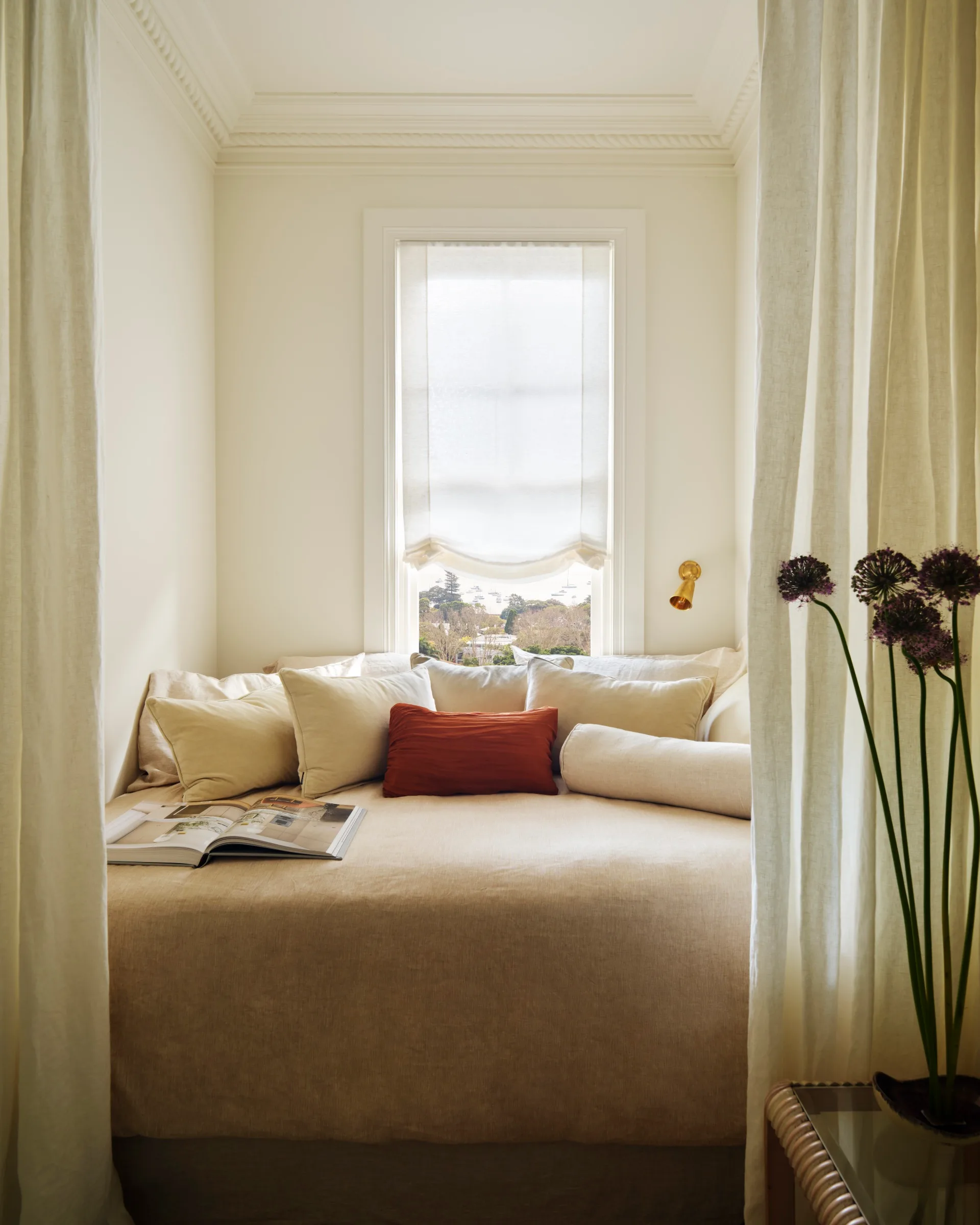 Cozy nook with beige bedding, pillows, open book, window view, and flowers beside.
