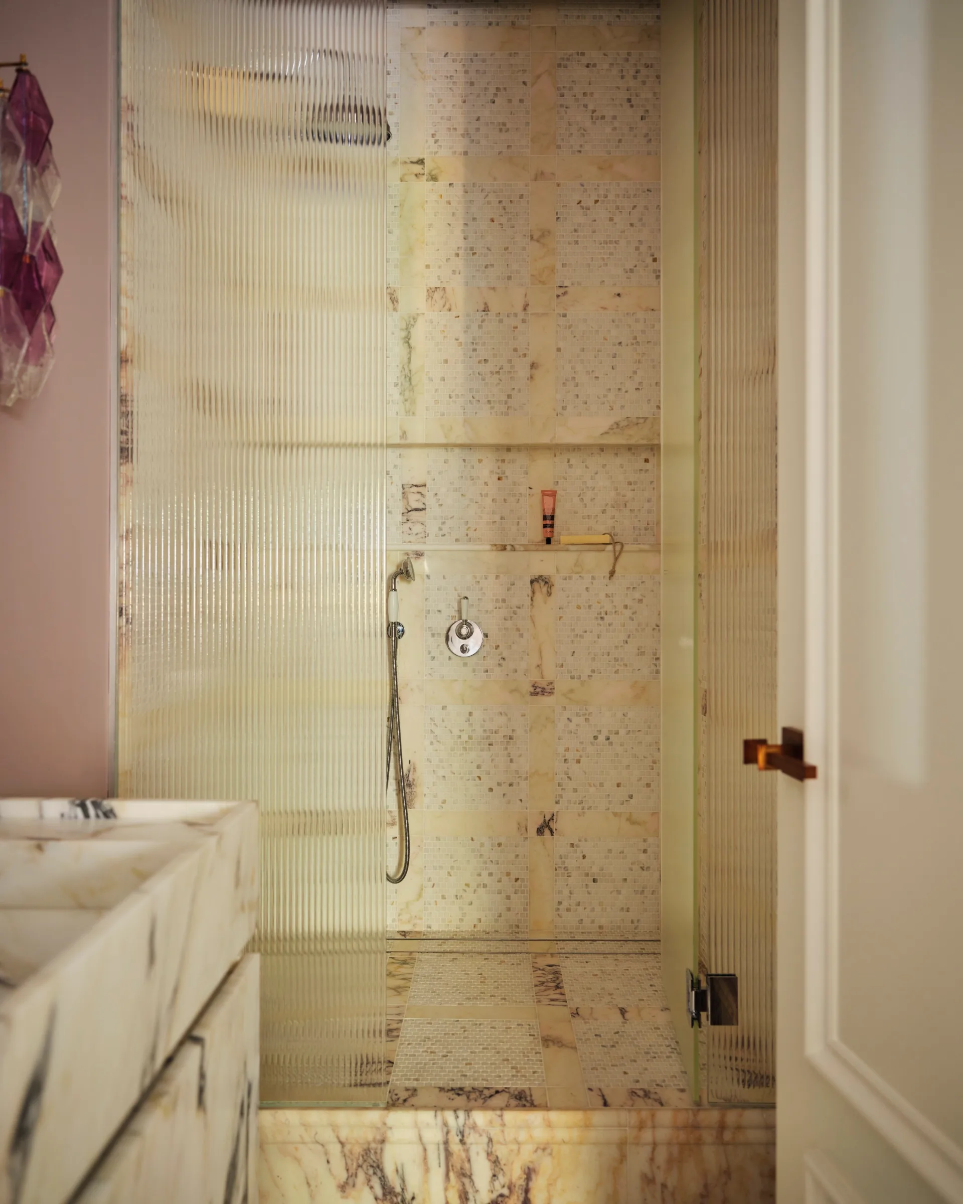 Luxurious marble shower with textured glass door, beige tiles, a handheld showerhead, and a pink toiletry bottle on a shelf.