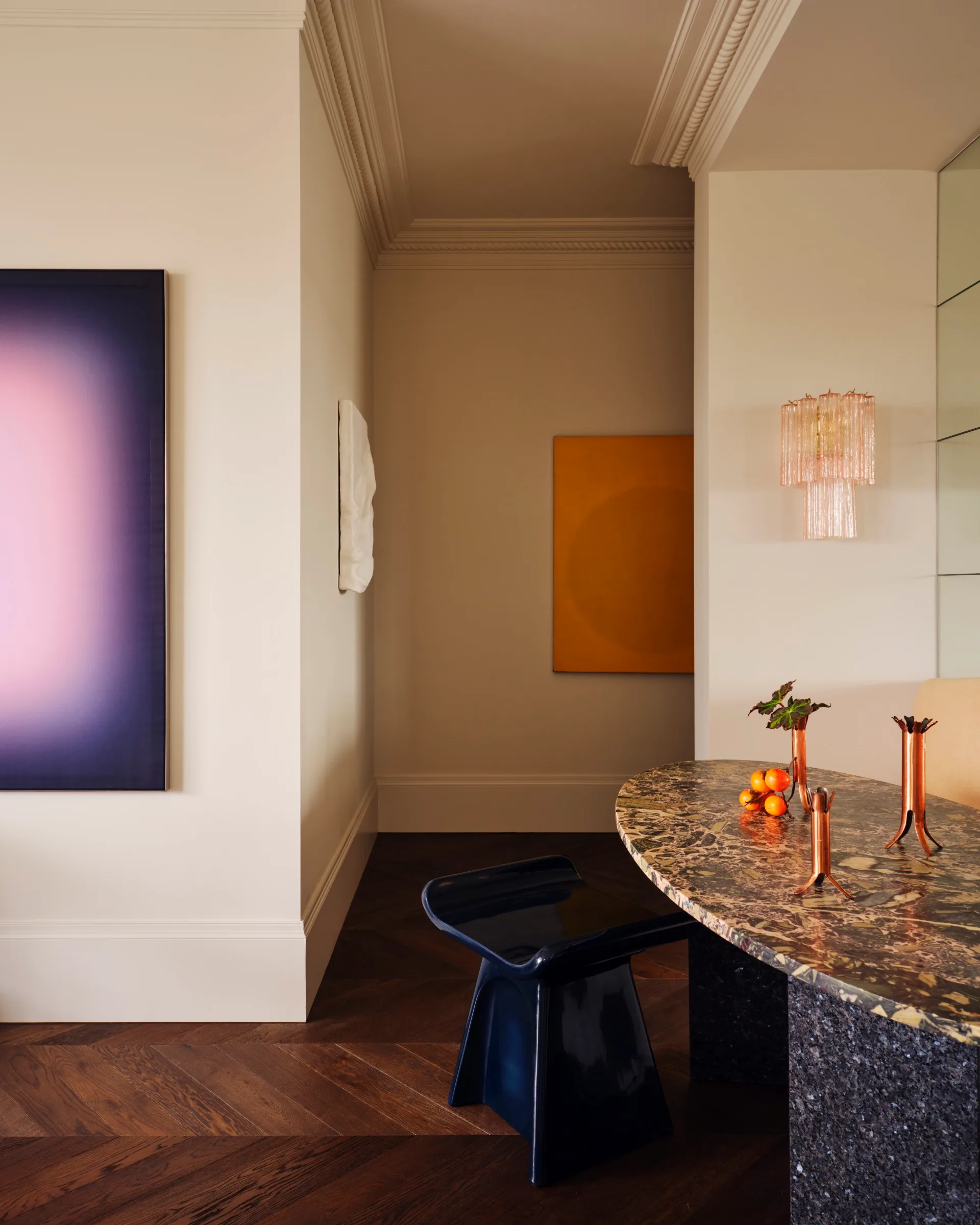 Elegant interior with abstract art, dark stool, marble table, pink light fixture, and decorative items on wooden flooring.