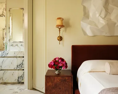 Bedroom with rust-colored bed, bedside table with red and pink roses, art piece on wall; marble bathroom visible.