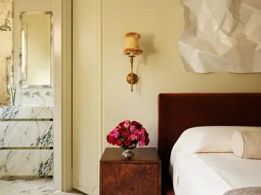Bedroom with rust-colored bed, bedside table with red and pink roses, art piece on wall; marble bathroom visible.