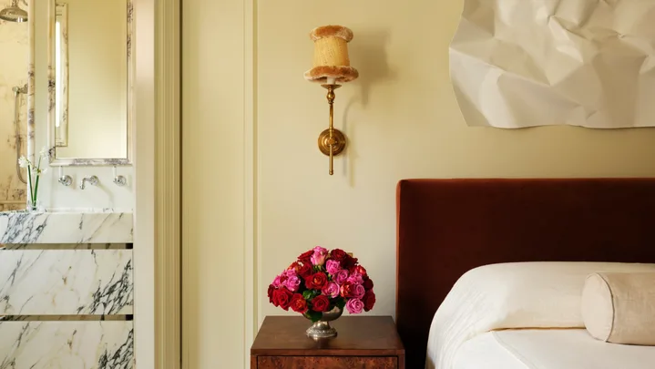 Bedroom with rust-colored bed, bedside table with red and pink roses, art piece on wall; marble bathroom visible.