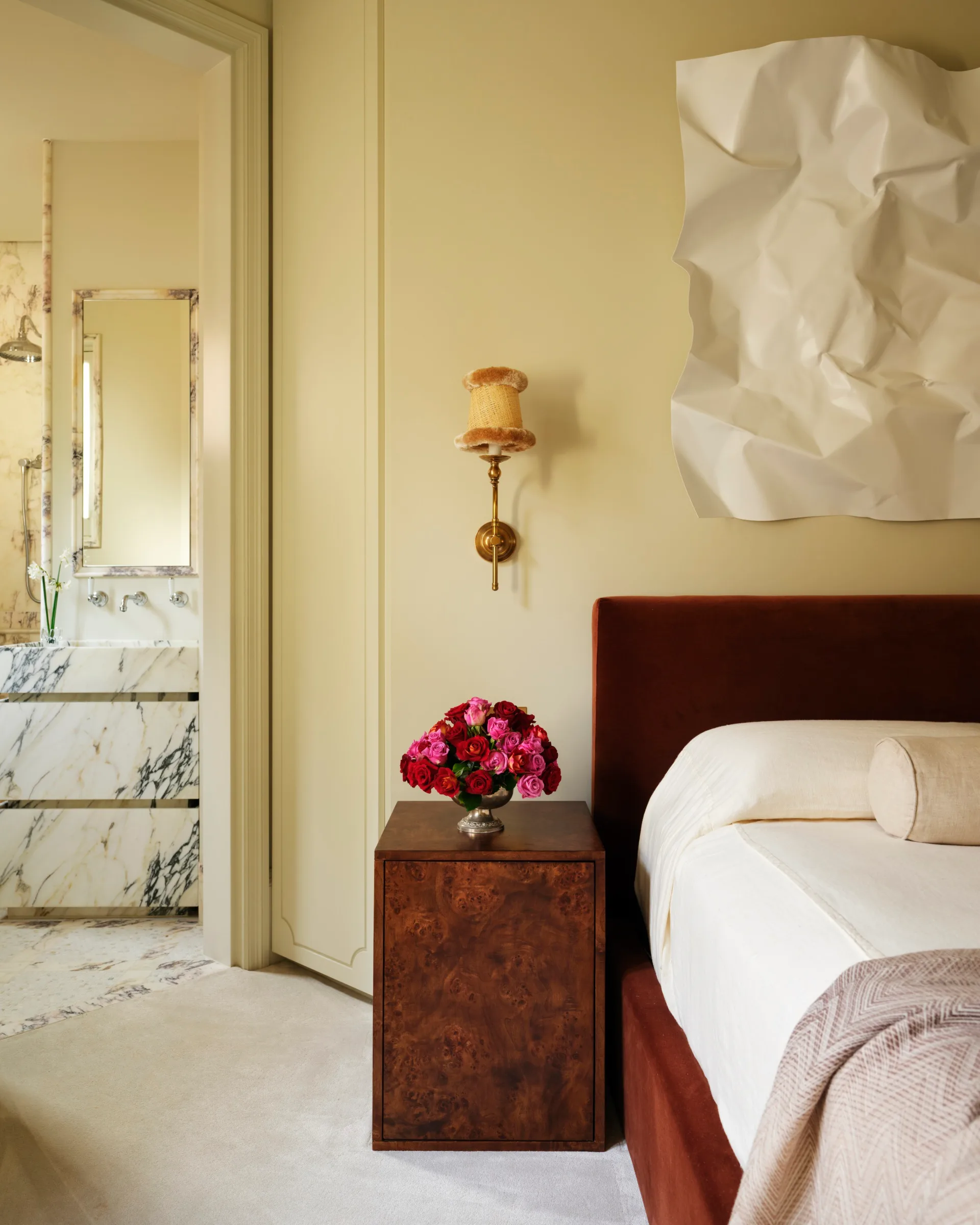 Bedroom with rust-colored bed, bedside table with red and pink roses, art piece on wall; marble bathroom visible.