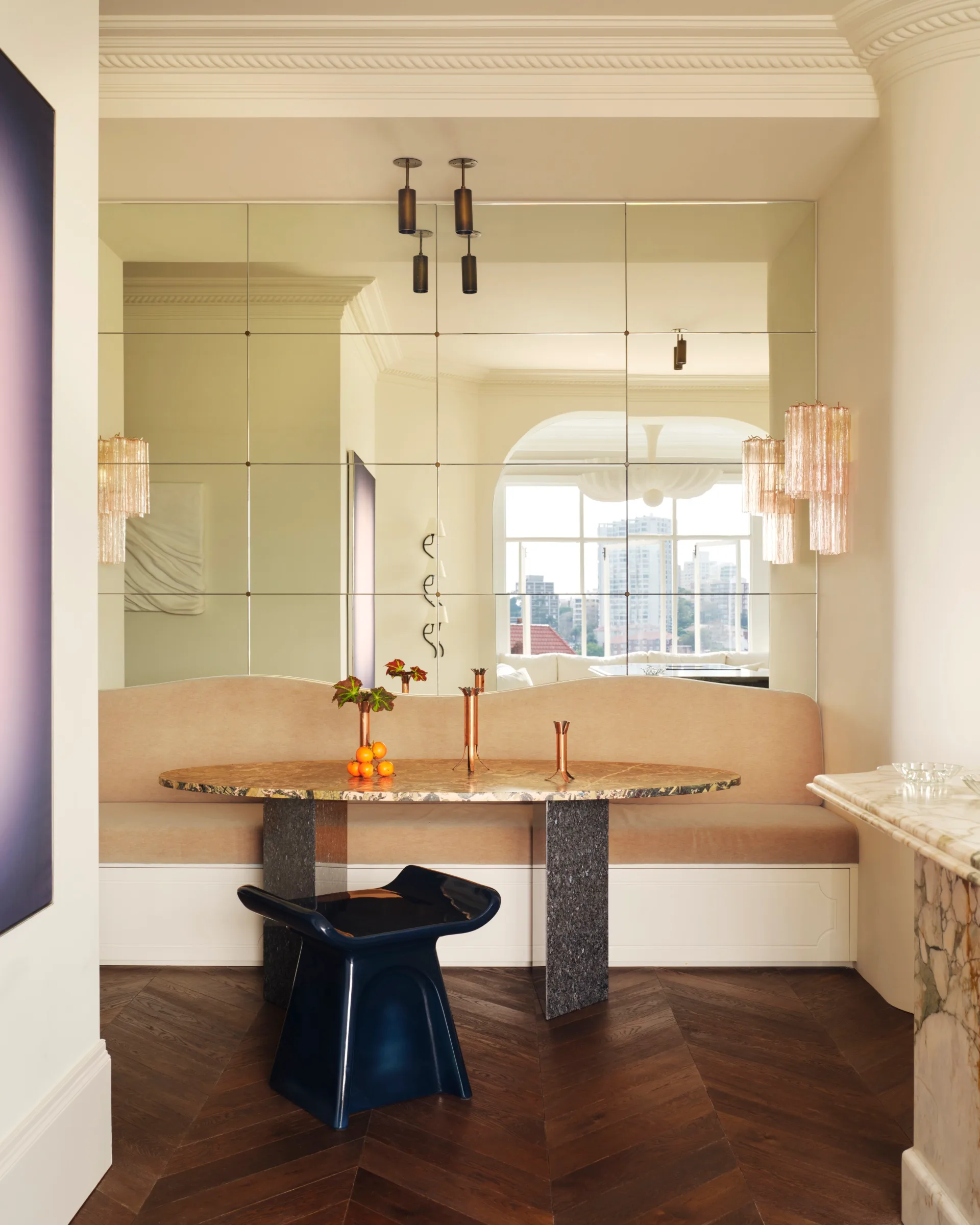 Bright modern dining nook with mirror wall, unique chairs, and city view. Decorative vases and chandelier accents.