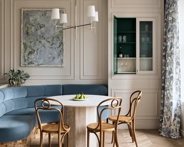 Stylish dining area with curved blue built-in banquette, abstract painting positioned in the middle of decorative panelling on the white walls, a round dining table, three burl wood dining chairs and floral drapes to the right. A cupboard door is open in built-joinery to the right of the dining table and banquette, revealing a small bar within.