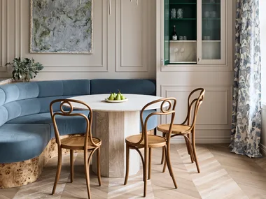 Stylish dining area with curved blue built-in banquette, abstract painting positioned in the middle of decorative panelling on the white walls, a round dining table, three burl wood dining chairs and floral drapes to the right. A cupboard door is open in built-joinery to the right of the dining table and banquette, revealing a small bar within.