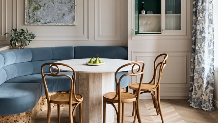 Stylish dining area with curved blue built-in banquette, abstract painting positioned in the middle of decorative panelling on the white walls, a round dining table, three burl wood dining chairs and floral drapes to the right. A cupboard door is open in built-joinery to the right of the dining table and banquette, revealing a small bar within.