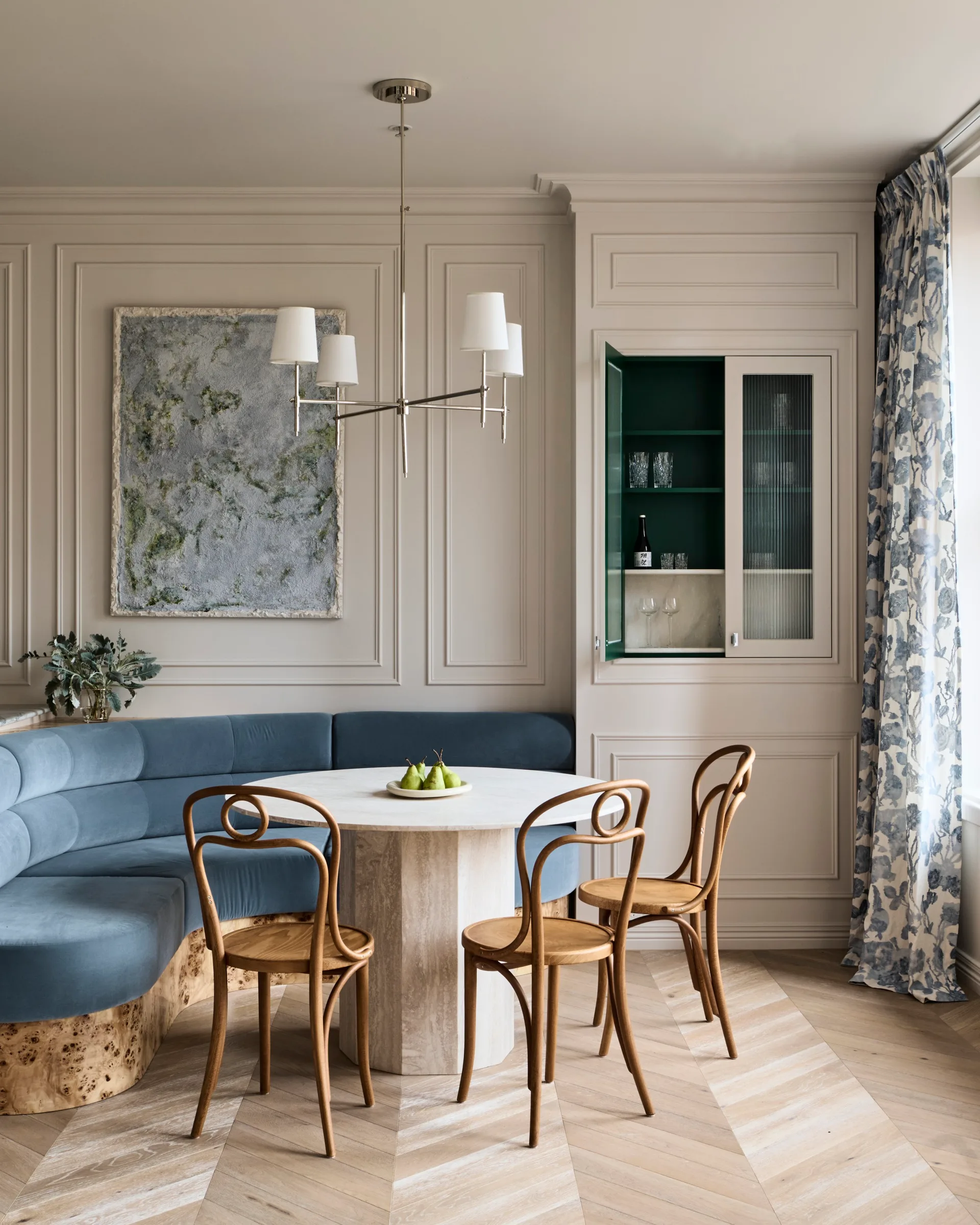 Stylish dining area with curved blue built-in banquette, abstract painting positioned in the middle of decorative panelling on the white walls, a round dining table, three burl wood dining chairs and floral drapes to the right. A cupboard door is open in built-joinery to the right of the dining table and banquette, revealing a small bar within.