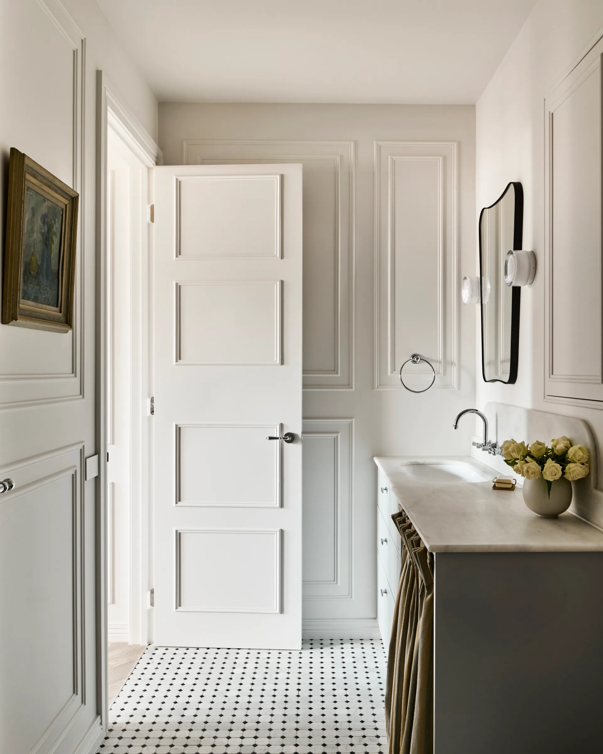 Elegant bathroom in a small apartment, with white paneled walls, an open door that is also white with decorative panelling, a mirror on the wall, and a vase of roses on the vanity countertop.